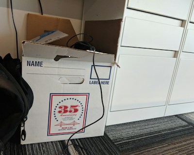 A cardboard box labeled for moving services is placed on a carpeted floor. The box has a design indicating a 35th anniversary. There is a cable draped over the box, and a backpack is partially visible next to it. Nearby, there are white drawers.