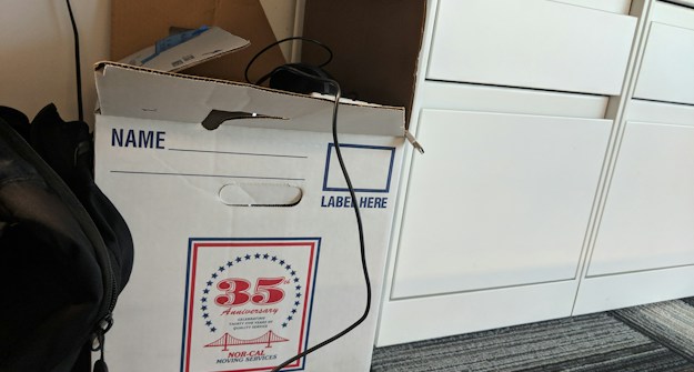 A cardboard box labeled for moving services is placed on a carpeted floor. The box has a design indicating a 35th anniversary. There is a cable draped over the box, and a backpack is partially visible next to it. Nearby, there are white drawers.