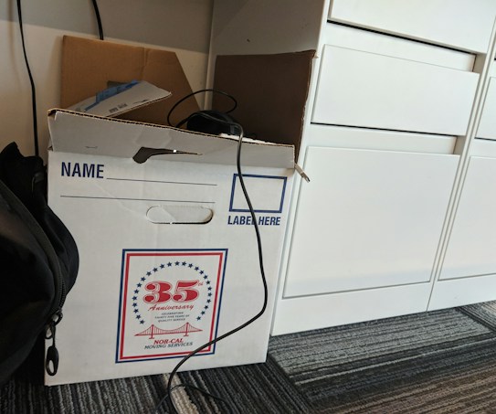 A cardboard box labeled for moving services is placed on a carpeted floor. The box has a design indicating a 35th anniversary. There is a cable draped over the box, and a backpack is partially visible next to it. Nearby, there are white drawers.