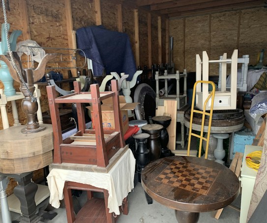 A cluttered storage area filled with various pieces of furniture and household items. Items include a checkerboard table, assorted chairs, stools, a hand truck, and other disassembled wooden furniture. The walls are unfinished, with exposed beams, creating a rustic backdrop.