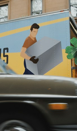 A vintage car is parked on a street with an urban backdrop, including brownstone buildings. In the background, a moving truck with a large graphic of a person carrying a box is visible. People are tending to a large potted plant, possibly relocating it from the truck or arranging it in its spot.