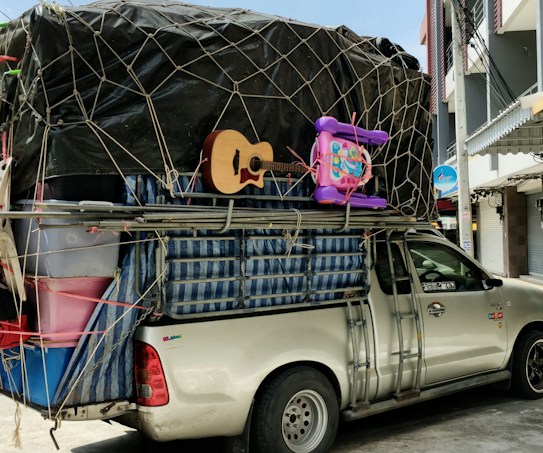 A pickup truck loaded with a variety of household items and furniture, secured with netting. The cargo includes a guitar, plastic bins, a mattress, and children's toys such as a tricycle and a baby walker. The truck is parked on a street adjacent to buildings and a tree, with some traditional lanterns hanging from a storefront.