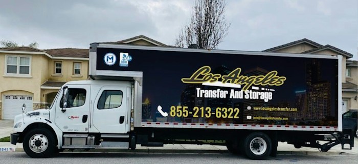 A large moving truck is parked on a residential street in front of a house with beige siding and white garage door. The truck is branded with 'Los Angeles Transfer And Storage,' featuring contact information and graphics of a city skyline. The sky is overcast, and the street is empty.