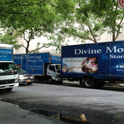 Several moving trucks from Divine Moving & Storage Ltd are parked along a street lined with trees. The trucks are large and blue, with company logos and contact information visible. A painting detail is incorporated into the truck design. The street has a sidewalk with some water reflecting the surroundings.