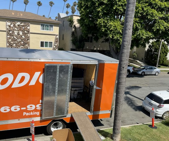 An orange moving truck is parked on a suburban street with its back door open. A ramp extends from the truck to the sidewalk, and there are traffic cones placed around the truck. Several cardboard boxes and items are visible inside the truck. Nearby, a tree provides shade, and apartment buildings line the street. There are also cars parked along the road.