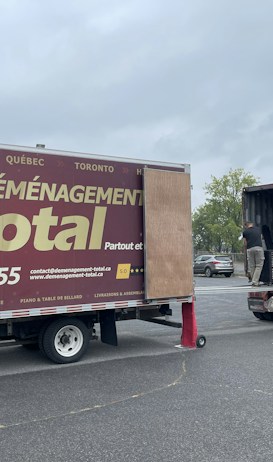 A moving truck with company branding is parked next to an open shipping container on a trailer. Two people are handling items near the container, while a car is parked in the background. The sky is overcast, and the ground appears to be asphalt.