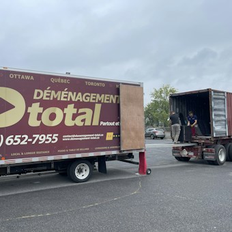 A moving truck with company branding is parked next to an open shipping container on a trailer. Two people are handling items near the container, while a car is parked in the background. The sky is overcast, and the ground appears to be asphalt.