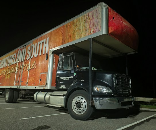 A large delivery truck is parked in a lot at night. The side of the truck features branding with the words 'Furnitureland South' and 'Let's Create Home' in bold, white lettering on a textured red and wood-patterned background. The front cab of the truck is black with silver accents and visible parking lines on the ground.