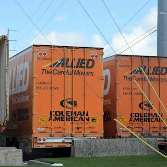 Two orange freight trailers are parked closely together, branded with the 'Allied' logo and the words 'The Careful Movers'. There are wires overhead and a partially visible open trailer to the left. The ground is concrete, and there are some trees and buildings in the background.