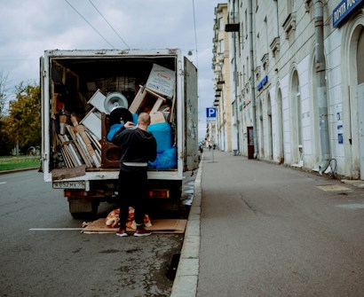 A person stands at the back of a moving truck on a city road, organizing a variety of items including boxes, a fan, and blue tarps. The truck is parked alongside a sidewalk bordered by a tall building with white stone walls and several business signs. The road appears empty except for parked cars and the scene is set under an overcast sky.
