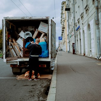 A person stands at the back of a moving truck on a city road, organizing a variety of items including boxes, a fan, and blue tarps. The truck is parked alongside a sidewalk bordered by a tall building with white stone walls and several business signs. The road appears empty except for parked cars and the scene is set under an overcast sky.