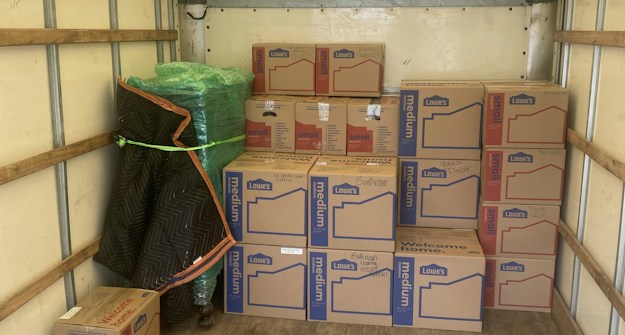 An empty truck interior with wooden floorboards is visible. Several cardboard boxes labeled 'Lowe's' are stacked neatly along the back of the truck. A green-wrapped item, possibly furniture, is secured at the side with a moving blanket partially covering it. There is a black and blue moving blanket and a green strap on the ground.