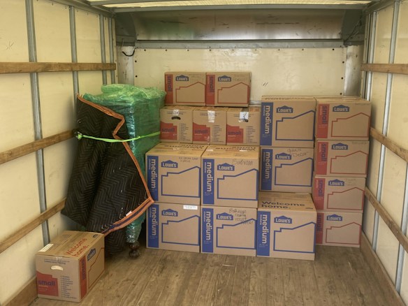 An empty truck interior with wooden floorboards is visible. Several cardboard boxes labeled 'Lowe's' are stacked neatly along the back of the truck. A green-wrapped item, possibly furniture, is secured at the side with a moving blanket partially covering it. There is a black and blue moving blanket and a green strap on the ground.